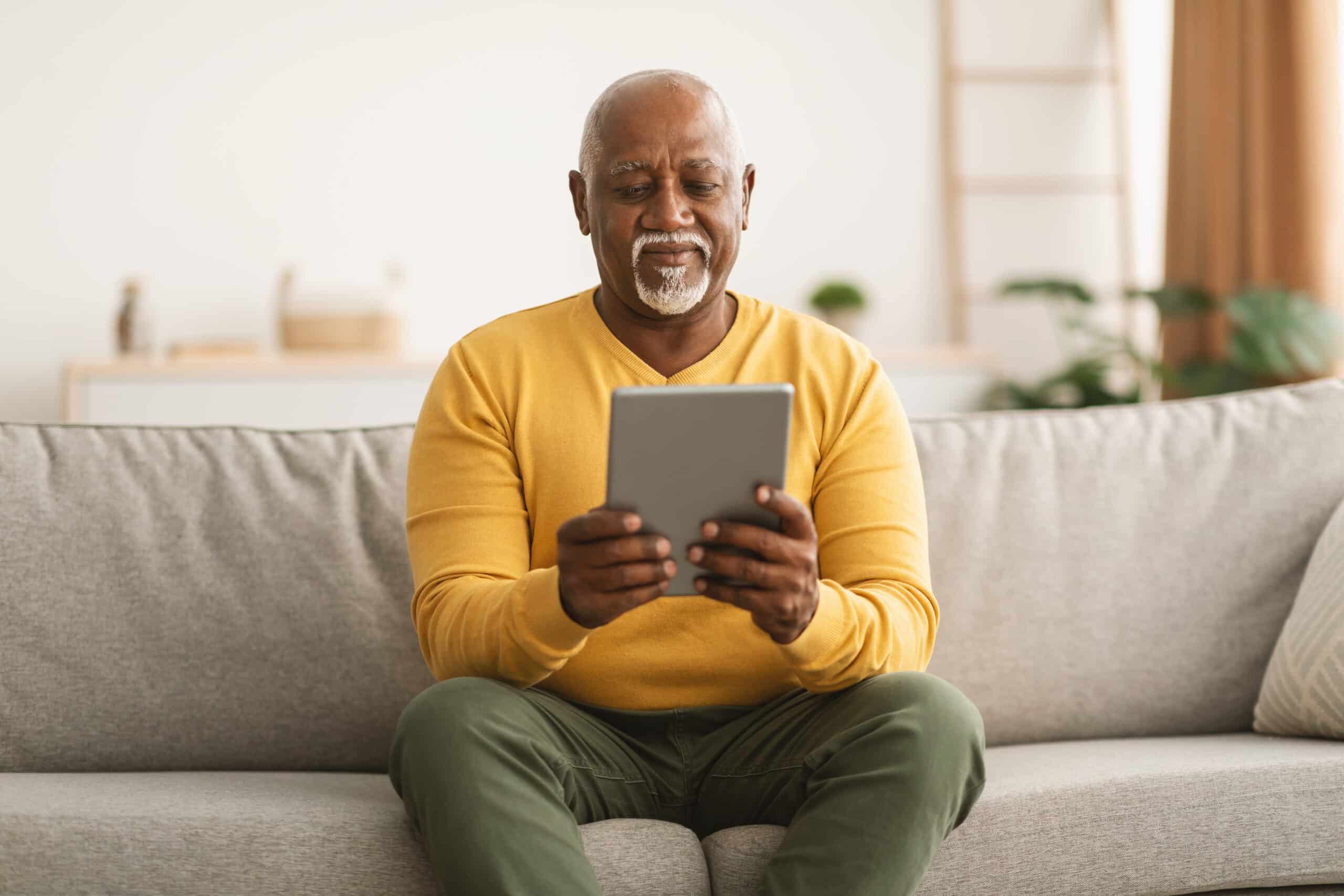 Senior African American Male Using Digital Tablet Reading Online Book And Browsing Internet Sitting On Sofa At Home. Mature Man Holding Computer Relaxing In Living Room. Older People And Gadgets